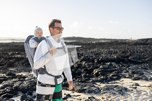 Image of Young father carrying his infant baby boy son in backpack on black rock volcanic beach on Lanzarote island, Spain. Family travel and winter vacation concept.