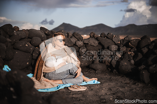 Image of Mother breast feeding his infant baby boy son on black sandy volcanic beach of Janubio on Lanzarote island, Spain, enjoing dramatic volcanic landscape on windy overcast day. Travel with kids concept
