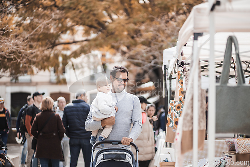 Image of Father walking carrying his infant baby boy child and pushing stroller in crowd of people wisiting sunday flea market in Malaga, Spain