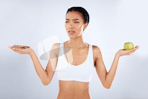 Image of Decide, studio and woman with a chocolate and apple to choose for a snack, craving or treat. Health, wellness and female model choosing candy or fruit for her healthy diet by a gray background.