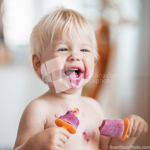 Image of Happy adorable infant baby boy child smiling while eating two frozen fruit popsicle ice creams in simmer.