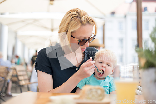 Image of Young caucasian blonde mother spoon feeding her little infant baby boy child outdoors on restaurant or cafe terrace in summer