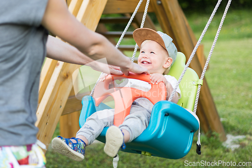 Image of Mother pushing her infant baby boy child on a swing on playground outdoors.