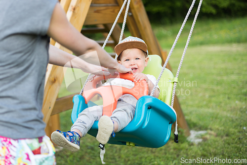 Image of Mother pushing her infant baby boy child on a swing on playground outdoors.