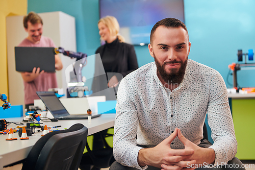 Image of A man sitting in a robotics laboratory while his colleagues in the background test new, cutting edge robotic inventions.