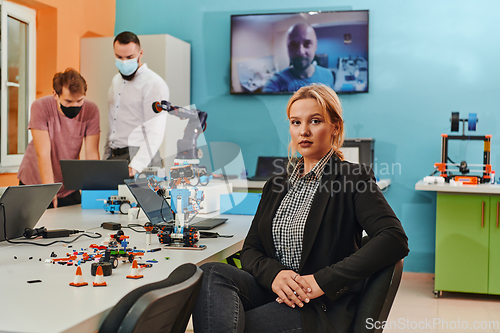 Image of A woman sitting in a laboratory and solving problems and analyzing the robot's verification. In the background, colleagues are talking at an online meeting