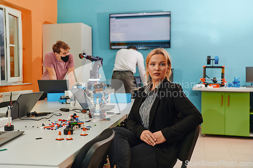 Image of A woman sitting in a laboratory and solving problems and analyzing the robot's verification. In the background, colleagues are talking at an online meeting