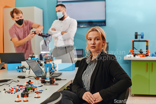 Image of A woman sitting in a laboratory and solving problems and analyzing the robot's verification. In the background, colleagues are talking at an online meeting