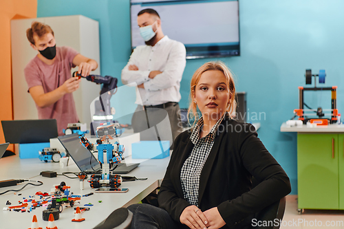 Image of A woman sitting in a laboratory and solving problems and analyzing the robot's verification. In the background, colleagues are talking at an online meeting