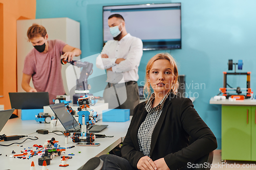 Image of A woman sitting in a laboratory and solving problems and analyzing the robot's verification. In the background, colleagues are talking at an online meeting