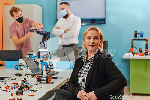 Image of A woman sitting in a laboratory and solving problems and analyzing the robot's verification. In the background, colleagues are talking at an online meeting