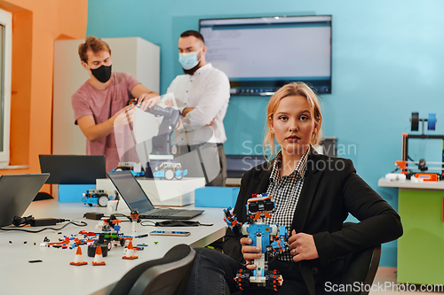 Image of A woman sitting in a laboratory and solving problems and analyzing the robot's verification. In the background, colleagues are talking at an online meeting