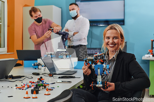 Image of A woman sitting in a laboratory and solving problems and analyzing the robot's verification. In the background, colleagues are talking at an online meeting