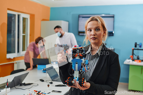 Image of A woman sitting in a laboratory and solving problems and analyzing the robot's verification. In the background, colleagues are talking at an online meeting