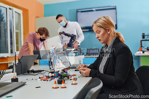 Image of A woman sitting in a laboratory and solving problems and analyzing the robot's verification. In the background, colleagues are talking at an online meeting