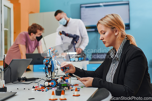 Image of A woman sitting in a laboratory and solving problems and analyzing the robot's verification. In the background, colleagues are talking at an online meeting