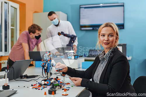 Image of A woman sitting in a laboratory and solving problems and analyzing the robot's verification. In the background, colleagues are talking at an online meeting