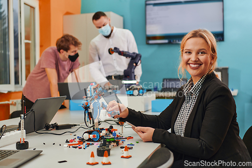 Image of A woman sitting in a laboratory and solving problems and analyzing the robot's verification. In the background, colleagues are talking at an online meeting