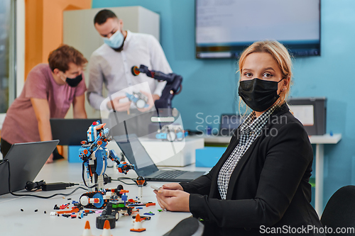 Image of A woman wearing a protective mask standing in a laboratory while her colleagues test a new robotic invention in the background.