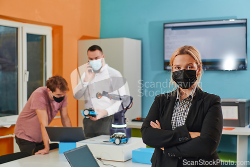 Image of A woman wearing a protective mask standing in a laboratory while her colleagues test a new robotic invention in the background.