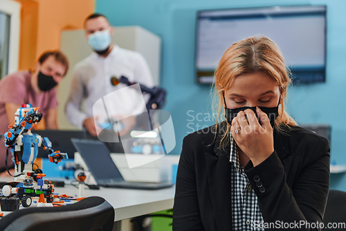 Image of A woman wearing a protective mask standing in a laboratory while her colleagues test a new robotic invention in the background.