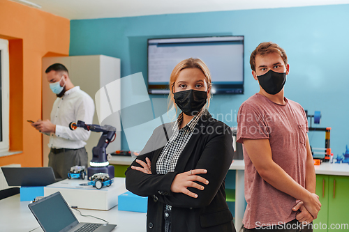 Image of A woman wearing a protective mask standing in a laboratory while her colleagues test a new robotic invention in the background.