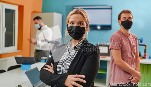 Image of A woman wearing a protective mask standing in a laboratory while her colleagues test a new robotic invention in the background.