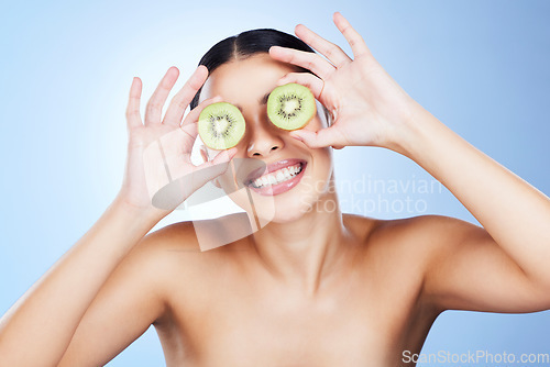 Image of Beauty, skincare and kiwi on eyes of woman feeling silly from fruit, wellness and vitamin c health. isolated, blue background and smile in studio of a young model happy about green cosmetic treatment