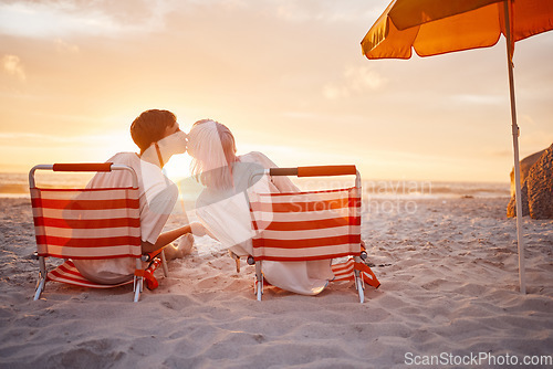 Image of Couple, kiss and beach chairs by a ocean sunset with love, happiness and care on vacation. Sea, sunshine and kissing of happy young people together sitting by the sand feeling relax outdoor