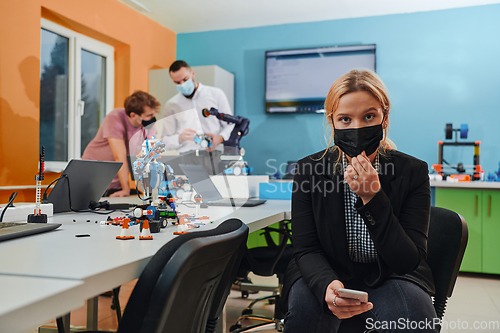 Image of A woman wearing a protective mask standing in a laboratory while her colleagues test a new robotic invention in the background.