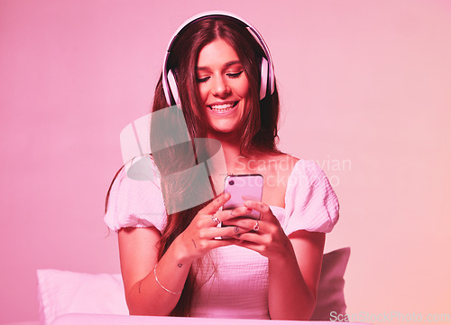 Image of Pink, light and music by woman in a bed with headphones, social media and streaming on wall background. Podcast, texting and girl relax in bedroom, happy and enjoying radio, audio or online playlist