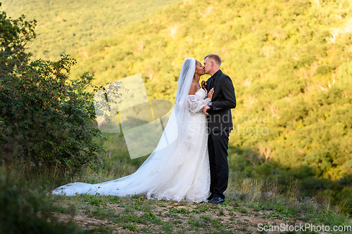 Image of Full-length portrait of the newlyweds against the backdrop of brightly lit foliage, the newlyweds are kissing