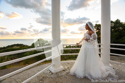 Image of A bride in a wedding dress in a gazebo with columns on the seashore looks towards the sea