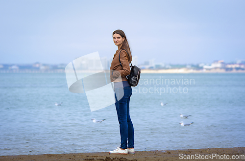 Image of A girl in autumn clothes with a backpack stands on the seashore, full-length portrait