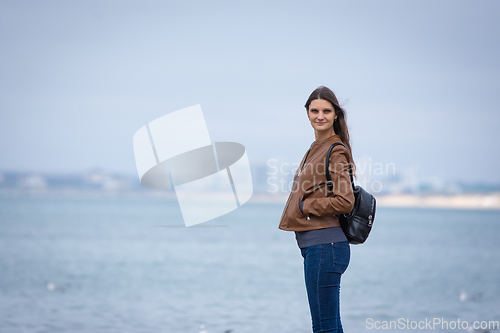 Image of A girl in autumn clothes with a backpack stands on the seashore