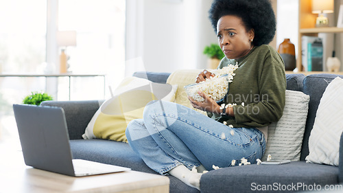 Image of African woman watching a horror movie on a laptop and eating popcorn while sitting on the couch at home. Terrified black afro female enjoying her online subscription with a variety of scary films
