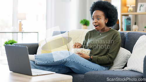 Image of Happy african american woman streaming online movies on a laptop while snacking on popcorn and relaxing on a sofa at home. Black female enjoying a comedy, eating, laughing and having fun on a weekend