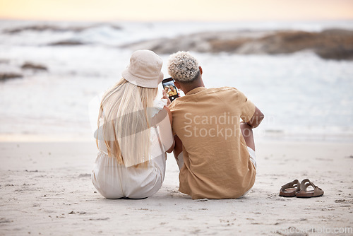 Image of Rear view of a young couple taking a break and taking a selfie with a smartphone at the beach during sunset. Caucasian female and her mixed race boyfriend relaxing and enjoying the view at the beach