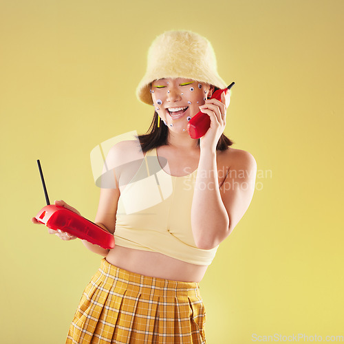 Image of Landline, fashion and woman on a call in a studio with art eyes stickers on her face with 90s aesthetic. Communication, beauty and happy Asian female with a telephone isolated by a yellow background.