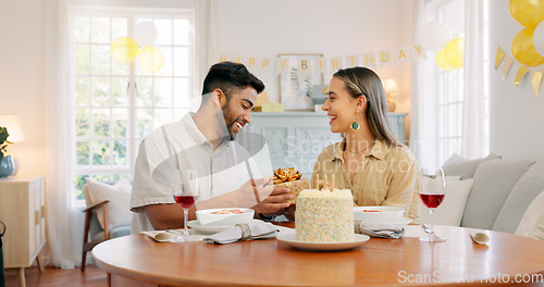 Image of Interracial, couple and celebrate for birthday with gift, being happy and relax together at party. Romance, man and woman with present, bonding and smile in home in living room for hug and embrace.