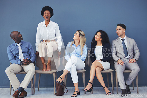 Image of Black woman, chair and smile with business people for interview with comic laugh, lobby or wall background. Group, we are hiring and waiting room with diversity, happy or opportunity at corporate job