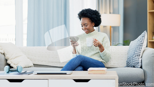 Image of Happy African American woman texting on a phone while relaxing on a sofa and drinking coffee at home. Smiling black woman browsing social media and laughing, planning on how to spend her free time