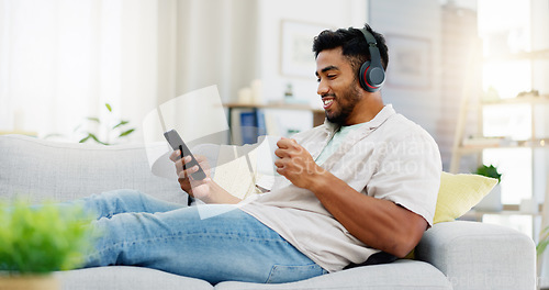 Image of Coffee, phone and happy man on sofa with headphones for social media, video streaming service and networking mobile app. Indian person relaxing on couch in his apartment on audio tech and smartphone