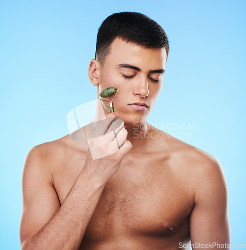 Image of Man, beauty and face with skin, roller for skincare and grooming with hygiene on blue background. Jade crystal, model and dermatology, cosmetic tools and treatment with facial massage in a studio