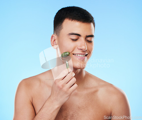 Image of Man, beauty and face with skincare, roller for skin and grooming with smile on blue background. Jade crystal, model and dermatology, cosmetic tools and treatment with facial massage in a studio