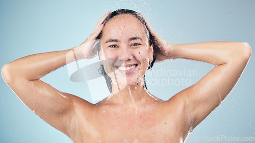 Image of Face, shower and woman smile washing hair in studio isolated on blue background. Water splash, hygiene and portrait of natural Asian model happy, cleaning or bathing for wellness, beauty and cosmetic