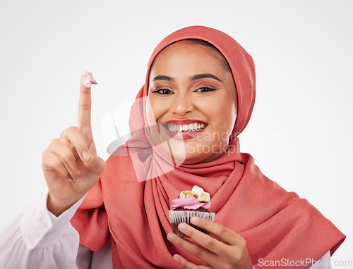 Image of Portrait, cupcake and icing on finger with a muslim woman in studio on a white background for dessert. Face, smile and food with a happy young islamic person eating a cake, candy or sugar snack