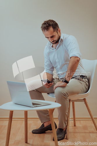 Image of A confident businessman sitting and using laptop and smartphone with a determined expression, while a beige background enhances the professional atmosphere, showcasing his productivity and expertise.