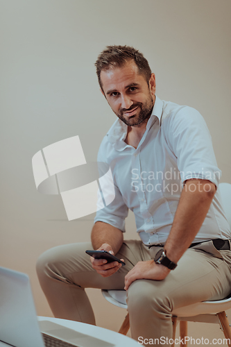 Image of A confident businessman sitting and using laptop and smartphone with a determined expression, while a beige background enhances the professional atmosphere, showcasing his productivity and expertise.