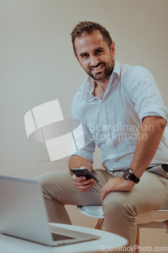 Image of A confident businessman sitting and using laptop and smartphone with a determined expression, while a beige background enhances the professional atmosphere, showcasing his productivity and expertise.